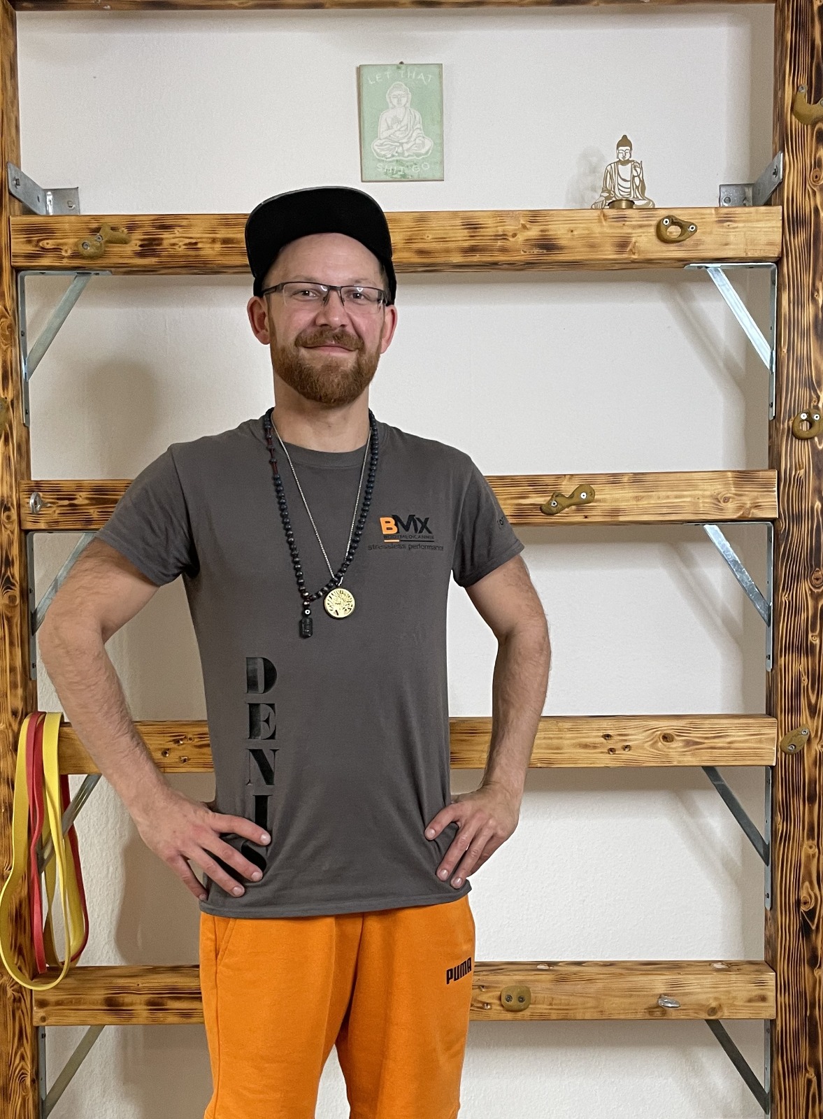 Ein Mann mit Brille, Bart und Kappe steht lächelnd vor einer Holz-Sprossenwand. Er trägt ein graues T-Shirt und orangefarbene Trainingshose.
