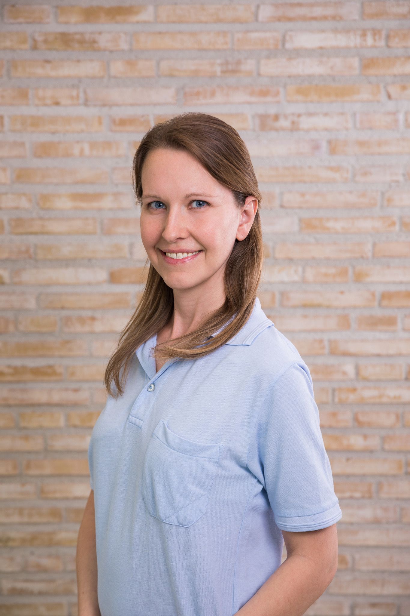 Eine freundliche Frau in hellblauem Polo-Shirt steht vor einer hellen Backsteinwand und lächelt in die Kamera. Physiotherapeutin: Michaela Lischka BSc MA
