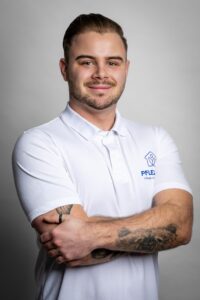 Ein junger Mann in weißem Polo-Shirt mit Logo des AS Pflegedienstes steht mit verschränkten Armen und lächelt in die Kamera.