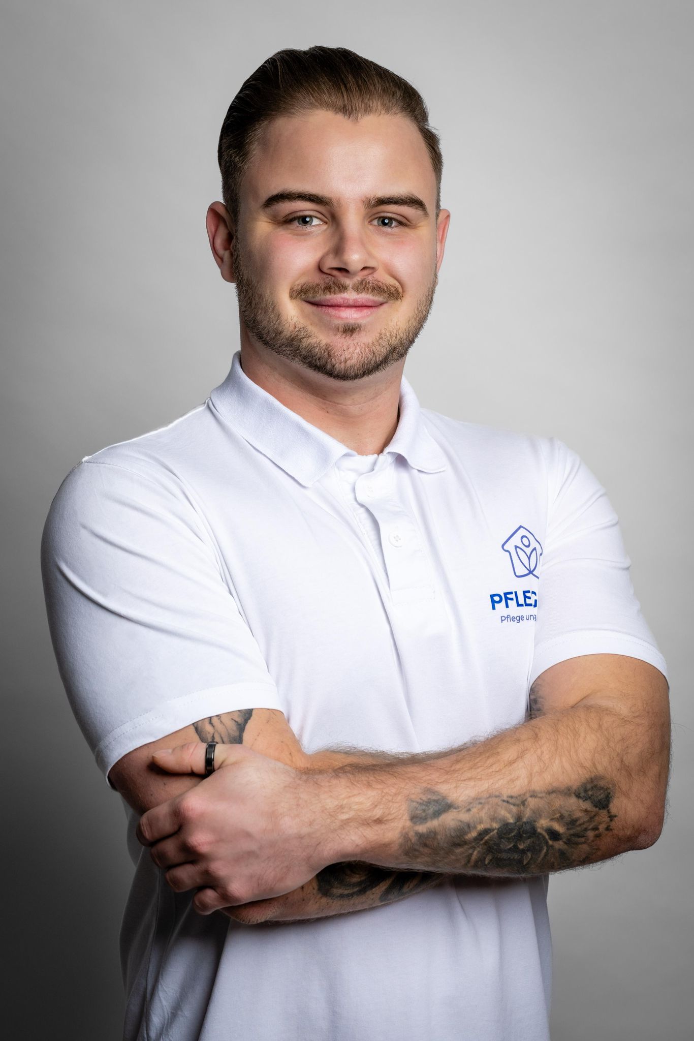Ein junger Mann in weißem Polo-Shirt mit Logo des AS Pflegedienstes steht mit verschränkten Armen und lächelt in die Kamera.