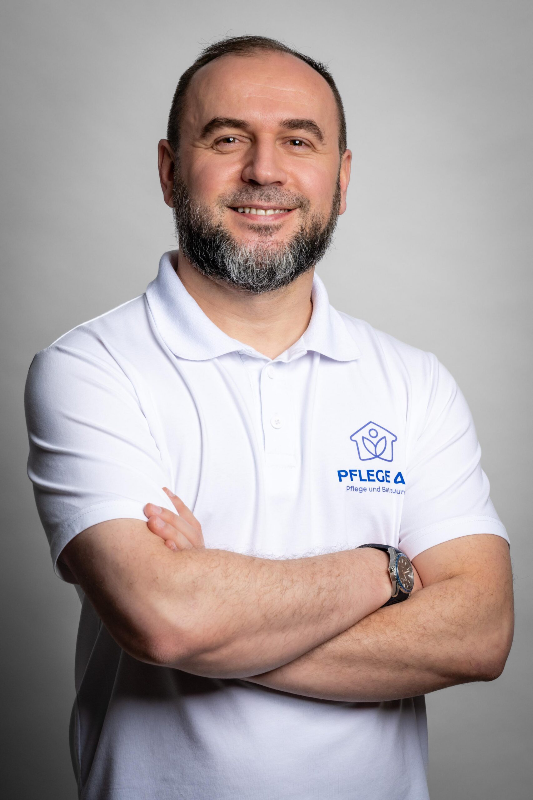 Ein Mann im weißen Polo-Shirt mit Logo des AS Pflegedienstes steht mit verschränkten Armen und lächelt freundlich in die Kamera.