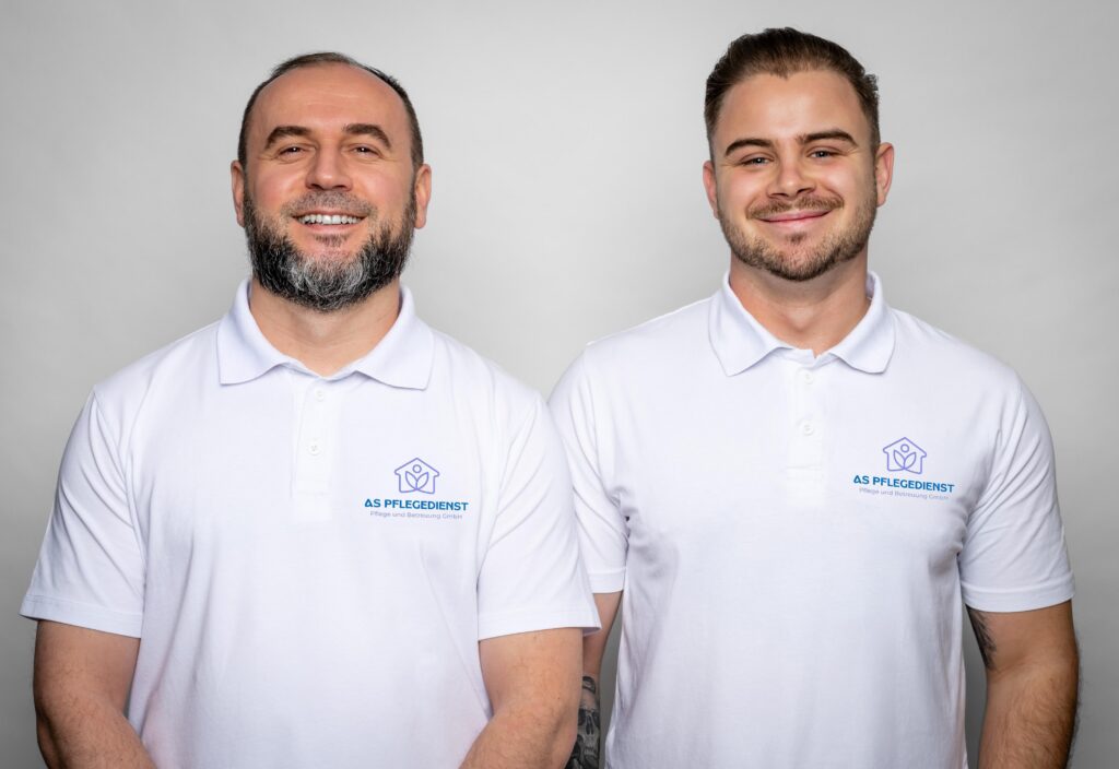 Zwei Mitarbeiter des AS Pflegedienstes tragen weiße Poloshirts mit dem Firmenlogo und lächeln in die Kamera.
